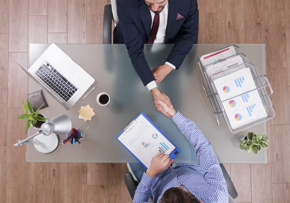 Top view of businessmen shaking hands during business negotiation in startup office discussing company strategy. Partners analyzing management profit planning investments meeting. Marketing documents